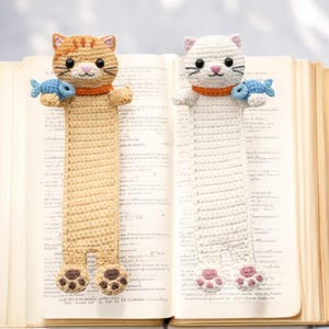 May include: Two crocheted cat bookmarks, one tan and one white, each featuring a cat head with black eyes, a pink nose, and an orange collar. Each cat holds a small blue fish in its paws and has visible paw pads on its feet at the bottom.