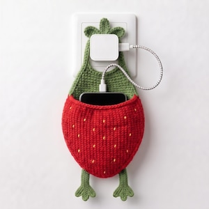 May include: A strawberry-shaped crochet phone holder in red with yellow seed accents and green leaves at the top and bottom. The holder hangs from a white wall power adapter plugged into an outlet, with a white charging cable connected to a phone.