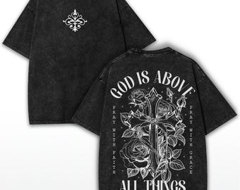 Pode incluir: T-shirt preto com lavagem ácida com um design floral branco na frente e um gráfico de cruz nas costas. O gráfico nas costas inclui o texto "GOD IS ABOVE ALL THINGS" e "PRAY WITH FAITH" e "PRAY WITH GRACE".