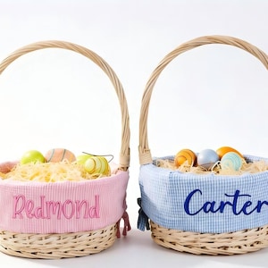 May include: Two woven Easter baskets with arched handles. One basket has a pink liner with the name "Redmond" embroidered in pink. The other has a blue gingham liner with "Carter" embroidered in blue. Both contain decorative eggs.