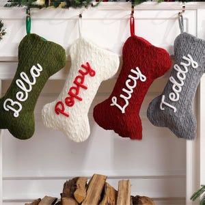 May include: Four bone-shaped Christmas stockings in green, white, red, and gray, each with a name embroidered in a contrasting color: Bella, Poppy, Lucy, and Teddy. The stockings are hanging from a white mantel.