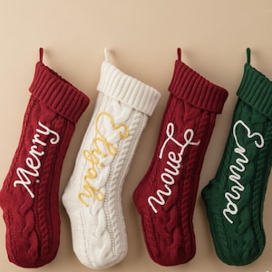 May include: Four cable-knit Christmas stockings with ribbed cuffs. Two are deep red with white embroidered names, one is cream with a yellow embroidered name, and one is dark green with a white embroidered name. Each stocking features a hanging loop.
