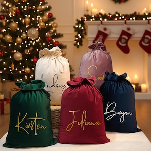 Pode incluir: Cinco sacos de presente de Natal de veludo em creme, verde, bordô, malva e azul marinho. Cada saco tem um fecho de cordão e um nome em escrita dourada ou branca: Lindsay, Kristen, Juliana, Sarah e Bryan. Uma árvore de Natal está ao fundo.
