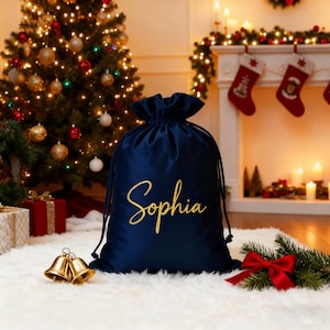 Puede incluir: Una bolsa de regalo de terciopelo azul marino con cordón, bordada con el nombre "Sophia" en letras doradas. La bolsa está sobre una alfombra blanca y esponjosa, con un árbol de Navidad y una chimenea al fondo.