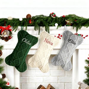 May include: Three cable-knit Christmas stockings in green, cream, and gray, each with a name embroidered on the front: "Chloe", "Nova", and "Ellie". The stockings hang from a white mantel decorated with greenery and ornaments.