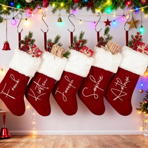 Puede incluir: Seis calcetines navideños rojos con puños de piel sintética blanca cuelgan de ganchos plateados. Cada calcetín está personalizado con un nombre en escritura blanca: Frank, Kristen, Joseph, Sophie y Arthur. Los calcetines están decorados con pequeños regalos y vegetación.