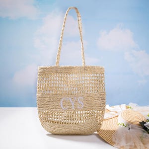 Puede incluir: Un bolso tote de paja tejida con forma rectangular y asas trenzadas. La bolsa presenta un diseño de tejido abierto y las iniciales "CYS" bordadas en blanco. Un sombrero de paja con adornos florales descansa cerca.