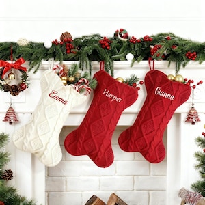 Puede incluir: Tres calcetines navideños cuelgan sobre una chimenea. Uno es blanco con el nombre "Emma" bordado en rojo, y los otros dos son rojos con los nombres "Harper" y "Gianna" bordados en blanco. Los calcetines están decorados con un patrón de punto trenzado.