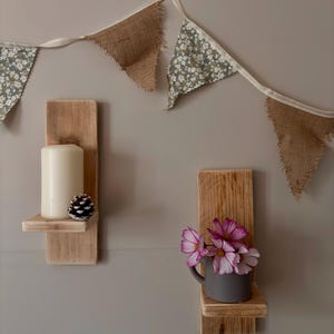 May include: Two wooden wall shelves, one with a white candle and pine cone, the other with a grey mug of pink and white flowers. A decorative banner with burlap and floral print triangles hangs above.