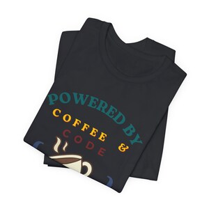 May include: A light gray heather t-shirt featuring the text "POWERED BY COFFEE & CODE" in teal and yellow, with a graphic of a smiling coffee cup inside curly brackets and a small ladybug, plus the text "fueled by caffeine and debugging".