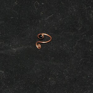 May include: A copper-colored nose ring with a small, round, black bead. The ring is crafted from a single piece of wire, with a spiral design on one end.