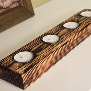 Może przedstawiać: Rustykalny drewniany świecznik z czterema świecami typu tealight. Drewno ma ciemne, spalone wykończenie, podkreślające usłojenie. Świece są białe, a uchwyt prostokątny. W tle znajduje się oprawiony obraz.
