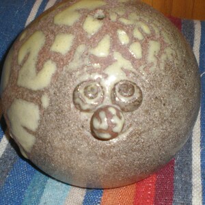 May include: A handmade ceramic sculpture with a quirky face design. The sculpture is mainly brown with cream-coloured accents and a textured surface. The face has three raised, circular elements for eyes and a nose, giving it a playful look.