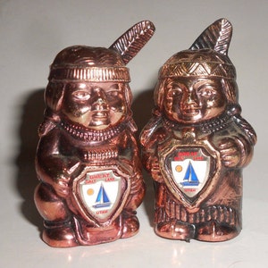 May include: A pair of copper-colored salt and pepper shakers shaped like Native American figures. Each figure is wearing a headdress with a feather and holding a shield with the words "Great Salt Lake, Utah".