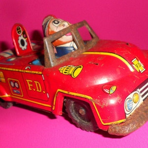 Puede incluir: Camión de bomberos de juguete rojo y amarillo con un conductor de dibujos animados. El camión tiene una campana en el capó y dice "F.D." en el lateral.