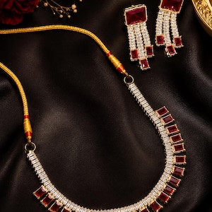 Könnte beinhalten: Eine Halskette und ein passendes Ohrringpaar mit rechteckigen roten Steinen und klaren Baguette-geschliffenen Kristallen in goldfarbenem Metall. Die Kette ist an einer gelben Kordel mit goldfarbenen Akzenten und einem runden Verschluss befestigt.