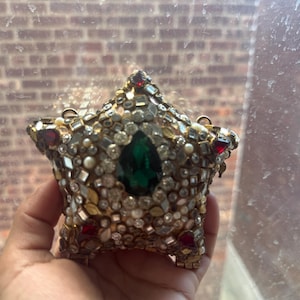 Pode incluir: Uma bolsa clutch em forma de estrela coberta por um mosaico intrincado de cristais transparentes, pequenas pérolas brancas e detalhes em metal dourado. Uma grande pedra preciosa verde esmeralda em forma de pera é o foco, com pedras vermelhas.