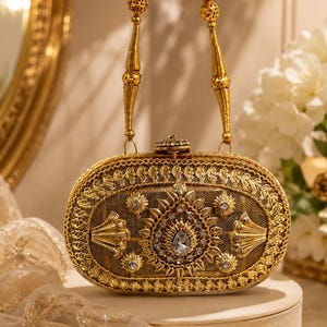 May include: An oval-shaped gold clutch bag featuring intricate metallic embroidery, a central teardrop-shaped crystal, and smaller embedded rhinestones. The bag includes a decorative gold chain handle with spherical filigree beads.