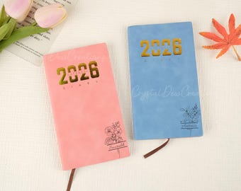 May include: Two hardcover diaries, one pink and one blue, with gold lettering that reads "2026 DIARY." Each diary has a floral design and a name. The pink diary has the name "Saurabh," and the blue diary has the name "Antonio."