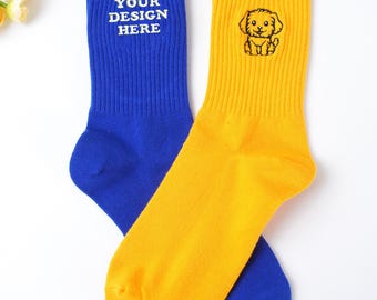 May include: Two colorful socks. One is blue with the text "YOUR DESIGN HERE" in white. The other is yellow with a small cartoon dog illustration. Both socks are ribbed and made of a soft material.