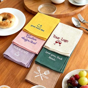 May include: A collection of six folded linen napkins in yellow, pink, light purple, cream, dark green, and tan. Each napkin features unique embroidered text and graphics, including phrases like good mood day, Custom design/text, NOOK CAFE, Your Logo, love you, Dad Noah, and FAST FOOD.