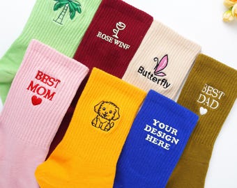 May include: A collection of colorful socks with embroidered designs. The socks feature various designs, including a palm tree, a wine glass, a butterfly, the words "BEST MOM" and "BEST DAD", a puppy, and the text "YOUR DESIGN HERE".