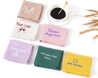 Puede incluir: Una colección de servilletas de lino dobladas en varios colores, cada una bordada con texto o diseños personalizados. Las servilletas incluyen texto como "FAST FOOD", "Your Logo", "Custom design/text", "NOOK CAFE", "Love you, Dad Noah" y "good mood day". Una taza de café en el fondo.