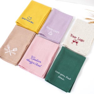 May include: An assortment of folded kitchen towels in shades like yellow, lilac, beige, pink, and green. Each towel is embroidered with text or a design, including phrases like "good mood day," "NOOK CAFE," and "FAST FOOD."