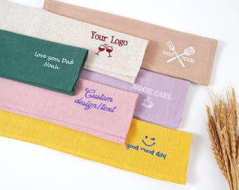 Puede incluir: Una colección de servilletas de lino coloridas con texto y diseños bordados. Las servilletas incluyen textos como "Your Logo", "Love you, Dad Noah", "Custom design/text", "NOOK CAFE" y "good mood day". Los colores incluyen verde, rosa, amarillo y beige.