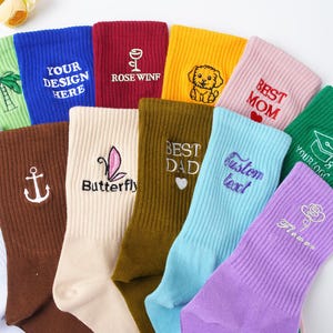May include: A collection of colorful ribbed socks with embroidered designs and text. The socks feature various designs, including a palm tree, anchor, butterfly, puppy, rose, and the words "YOUR DESIGN HERE," "ROSE WINF," "BEST MOM," "BEST DAD," "Custom text," and "Flower."