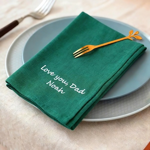 May include: An emerald green napkin with the embroidered text "Love you, Dad Noah" sits on a blue plate. A gold-coloured fork with a leaf design rests on the napkin. A silver fork is visible in the background.