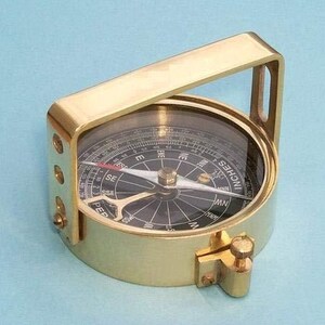 May include: A brass compass with a handle and a clear glass top. The compass face is black with white markings and a needle pointing north. The compass has a screw to lock the needle in place. The word "INCHES" is visible on the compass.