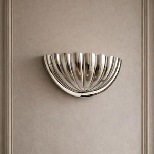 Könnte beinhalten: Eine halbkreisförmige Wandleuchte mit polierter silberner Oberfläche und einem geriffelten, muschelartigen Design. Die Leuchte weist vertikale Rillen auf, die von der zentralen Basis ausgehen und eine reflektierende, geschwungene Form bilden.