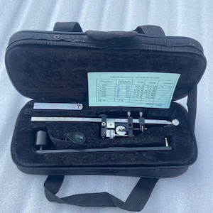 Op de afbeelding: Een open zwarte koffer met een meetinstrument en accessoires. De koffer bevat een precisiegereedschap met een wijzerplaat, een liniaal en een kleine groene kaart met tekst. De koffer heeft een handvat.