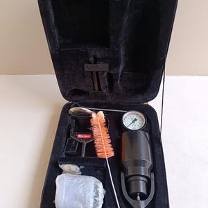 Puede incluir: Maleta negra abierta con ribete rojo, que contiene un medidor de humedad y accesorios. La maleta contiene un medidor de humedad negro, un cepillo de limpieza y otras herramientas. El medidor de humedad tiene un indicador con una aguja y un cuerpo negro.