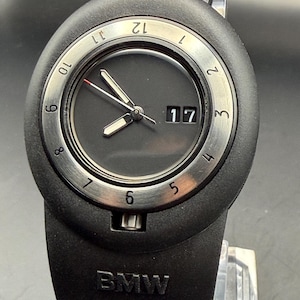 May include: Black wristwatch with a silver bezel and a black dial. The watch features a date display and luminous hands. The watch band is black and has the BMW logo. The watch is a stylish accessory.
