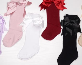 May include: A collection of knee-high socks in various colors, including gray, pink, white, red, black, and beige. Each sock features a large satin bow at the top in a matching or contrasting color. The socks are displayed on a white surface.