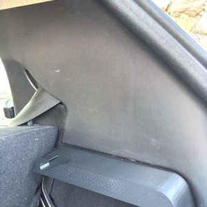 Op de afbeelding: Een close-up van het interieur van een auto, met zwarte en grijze panelen. De afbeelding toont een veiligheidsgordel, een plank en gestructureerde oppervlakken. De materialen lijken stof en plastic te zijn.