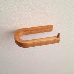 Può includere: Un portarotolo per carta igienica in legno marrone chiaro a forma di U con una finitura liscia e un piccolo piolo cilindrico all'estremità del braccio inferiore. Il supporto presenta una venatura del legno visibile ed è montato orizzontalmente.