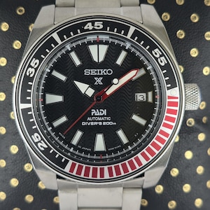 Può includere: Orologio subacqueo automatico Seiko PADI in acciaio inossidabile con quadrante nero e lunetta girevole rossa e nera. L'orologio presenta indici bianchi, visualizzazione della data e cinturino in acciaio inossidabile. Resistente all'acqua fino a 200m.