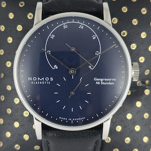 Puede incluir: Un reloj de pulsera Nomos Glashütte con esfera azul oscuro, manecillas plateadas y correa de cuero negro. La esfera presenta un indicador de reserva de marcha con el texto Gangreserve 48 Stunden, un subesfera de segundos y la marca Nomos Glashütte.