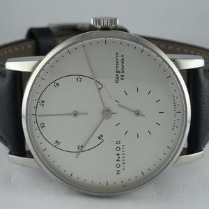 Puede incluir: Un reloj de pulsera Nomos Glashütte con esfera de color blanco plateado, índices negros finos y correa de cuero negro. La esfera presenta el texto "NOMOS GLASHÜTTE", "Gangreserve 48 Stunden" y "MADE IN GERMANY", con un indicador de reserva.