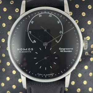 Puede incluir: Un reloj de pulsera Nomos Glashütte con caja redonda plateada y esfera negra. La esfera presenta marcadores de hora blancos, manecillas finas, una subesfera para los segundos y un indicador de reserva de marcha con la etiqueta Gangreserve 48 Stunden.