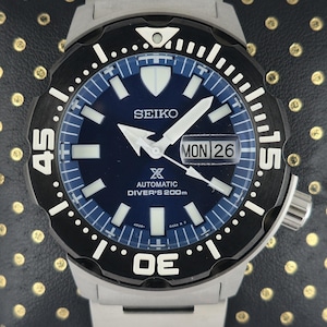 Op de afbeelding: Een roestvrijstalen Seiko automatische duikershorloge met een blauwe wijzerplaat en een zwarte draaibare bezel. Het horloge heeft witte uurmarkeringen, wijzers en een dag-datumweergave. Het horloge is waterdicht tot 200m.