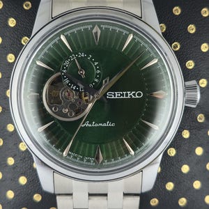 Może przedstawiać: Srebrny automatyczny zegarek Seiko z zieloną tarczą. Zegarek posiada widoczny wychwyt, srebrne wskazówki i wskaźnik daty. Pasek zegarka wykonany jest ze srebrnych ogniw. Na tarczy zegarka widnieją napisy "SEIKO" i "Automatic".