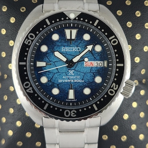 Op de afbeelding: Een roestvrijstalen Seiko automatische duikershorloge met een blauwe textuur wijzerplaat. Het horloge heeft een zwarte draaibare bezel, lichtgevende wijzers en markeringen en een dag-datum weergave. De band is van roestvrij staal.