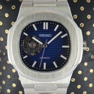 May include: A silver Seiko automatic wristwatch with a blue dial. The watch has a visible movement and a stainless steel strap. The face has white hour markers and hands. The watch is displayed on a black surface.