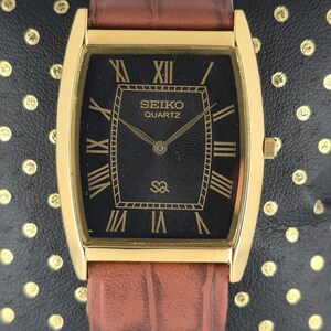 May include: A rectangular gold-toned Seiko Quartz watch with a black face and Roman numeral hour markers. The watch has a brown leather strap. The watch face displays the Seiko Quartz logo and a small symbol. The watch is displayed on a black surface.