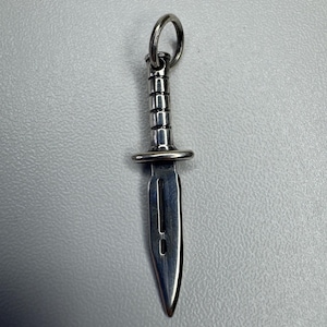 Puede incluir: Colgante de daga plateada con un anillo para sujetar. El mango es detallado y la hoja puntiaguda. El colgante probablemente está hecho de acero inoxidable o un metal similar. Un pequeño artículo decorativo.