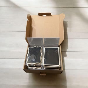May include: An open cardboard box containing several stacks of small black chalkboards with wooden frames. Some chalkboards have a decorative, scalloped edge. The chalkboards are packaged in clear plastic sleeves.
